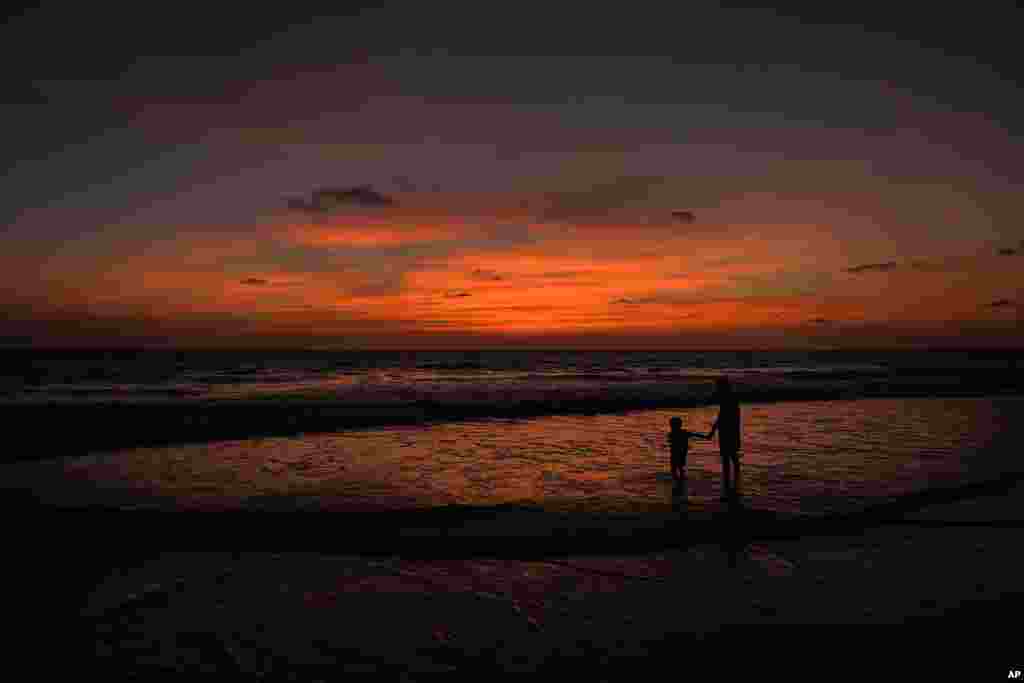 A boy holds the hand of his father as they watch the sun set at a beach in Goa, India.