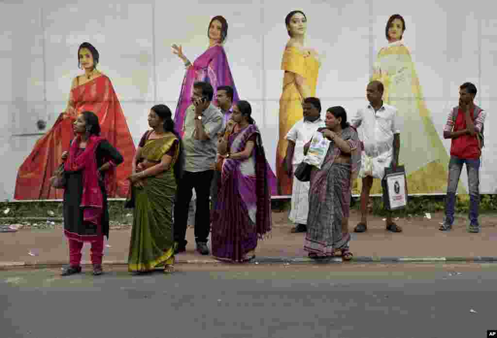 Indians returning after the day's work await bus transport standing on a pavement next to an advertisement hoarding of a clothing store in Thiruvananthapuram, Kerala state, India.