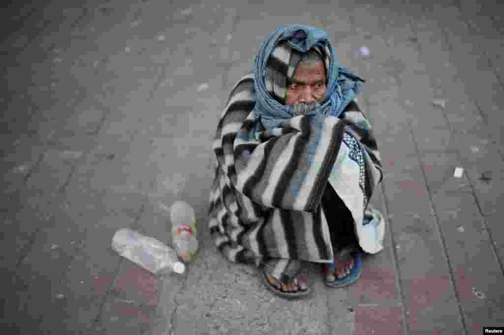 A man is covered in a quilt to keep himself warm as he sits on a pavement on a cold winter morning in New Delhi, India.