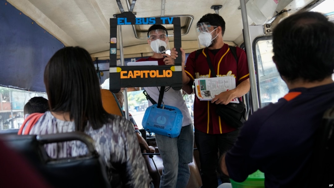 Juan Pablo Lares, right, holds a cardboard frame in front of his associate Maximiliano Bruzual who reads their newscast "El Bus TV Capitolio" to commuters on a bus in Caracas, Venezuela. (AP Photo/Ariana Cubillos)